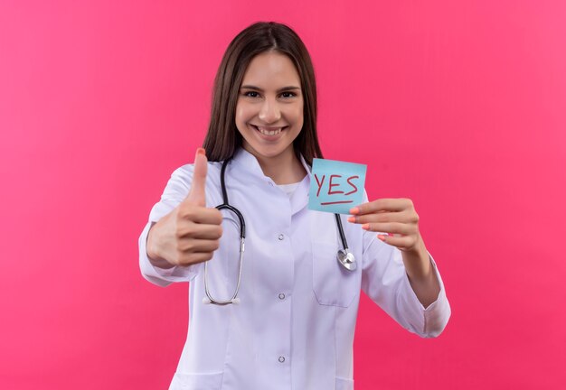lächelndes junges Doktormädchen, das medizinisches Kleid des Stethoskops hält Papier hält, markiert ihren Daumen oben auf isolierter rosa Wand