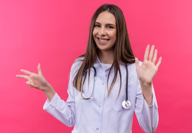 Lächelndes junges Doktormädchen, das medizinisches Kleid des Stethoskops trägt, das verschiedene Zahlen auf lokalisiertem rosa Hintergrund zeigt