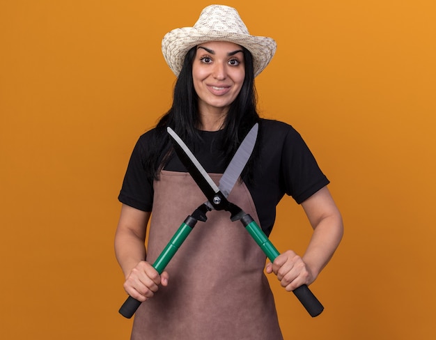 Lächelndes junges Gärtnermädchen in Uniform und Hut mit Heckenschere mit Blick auf die Vorderseite isoliert auf oranger Wand