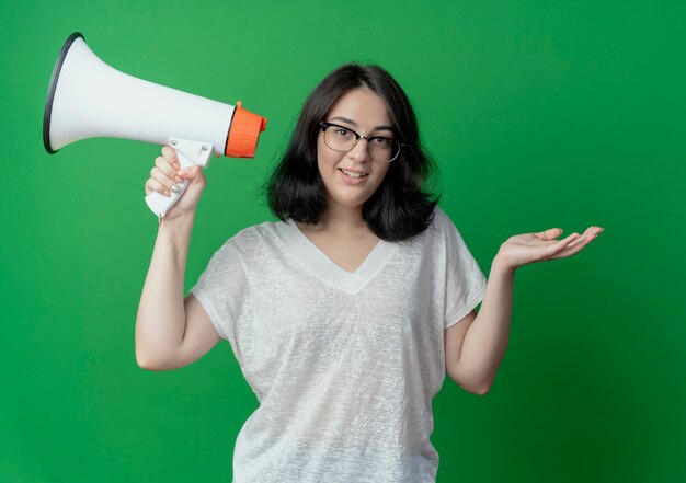 Lächelndes junges hübsches kaukasisches Mädchen, das die Brille hält, die Sprecher hält und leere Hand lokalisiert auf grünem Hintergrund zeigt