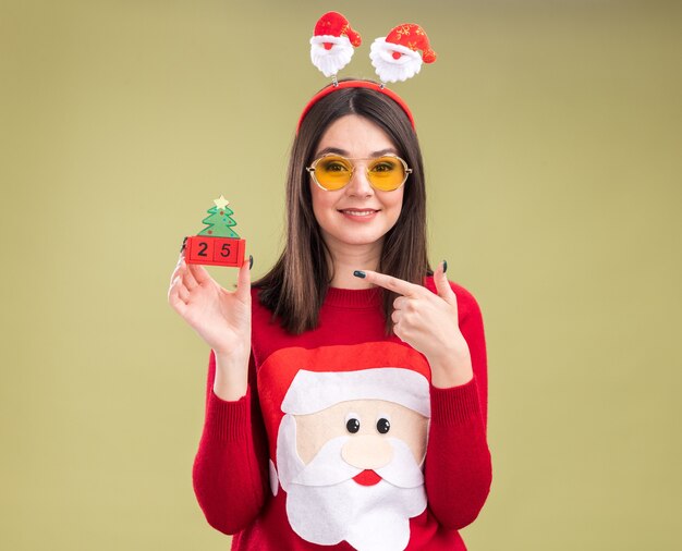 Lächelndes junges hübsches kaukasisches Mädchen, das Weihnachtsmann-Pullover und Stirnband mit Brille hält, die auf Weihnachtsbaumspielzeug mit Datum hält und zeigt