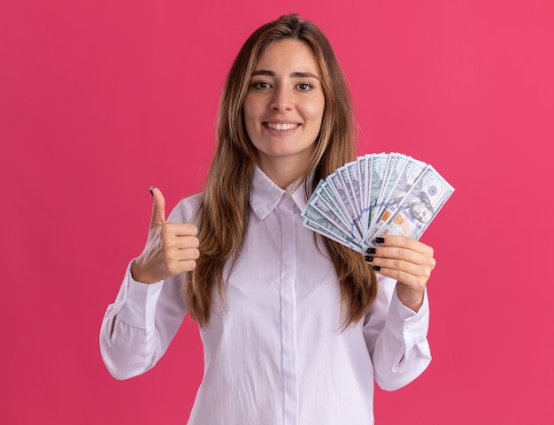 Lächelndes junges hübsches kaukasisches Mädchen Daumen hoch und hält Geld auf rosa