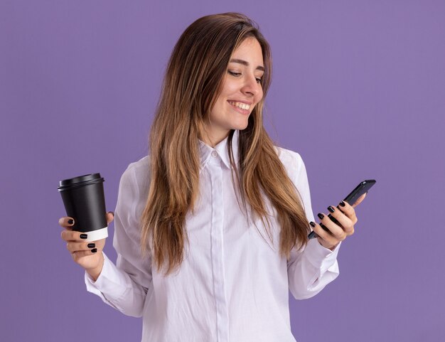 Lächelndes junges hübsches kaukasisches Mädchen hält Pappbecher und schaut auf Telefon auf lila