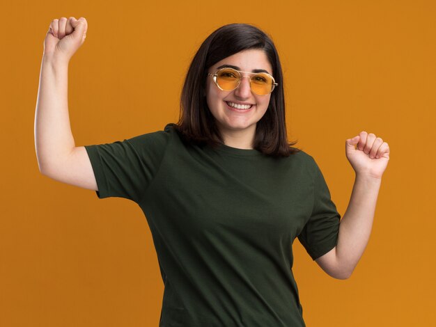 Lächelndes junges hübsches kaukasisches Mädchen in der Sonnenbrille steht mit erhobenen Fäusten auf Orange