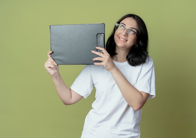 Lächelndes junges hübsches kaukasisches Mädchen mit Brille, das die Zwischenablage auf olivgrünem Grünraum hält