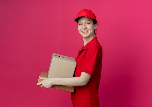 Lächelndes junges hübsches Liefermädchen, das rote Uniform und Kappe trägt, die in der Profilansicht hält, die Kartonschachtel und Pizzapaket lokalisiert auf purpurrotem Hintergrund mit Kopienraum hält