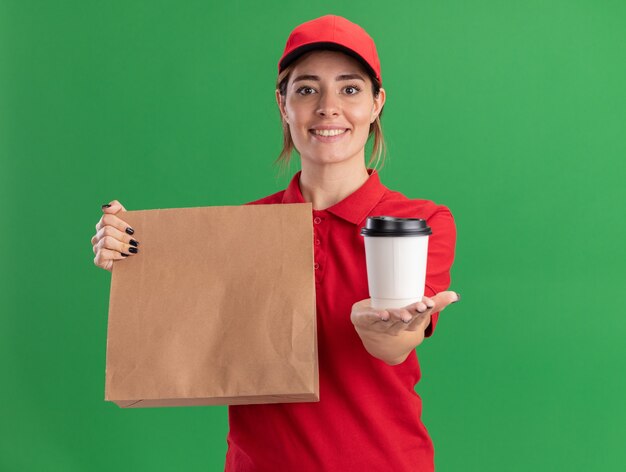 Lächelndes junges hübsches Liefermädchen in der Uniform, die Papierpaket und Pappbecher auf Grün hält