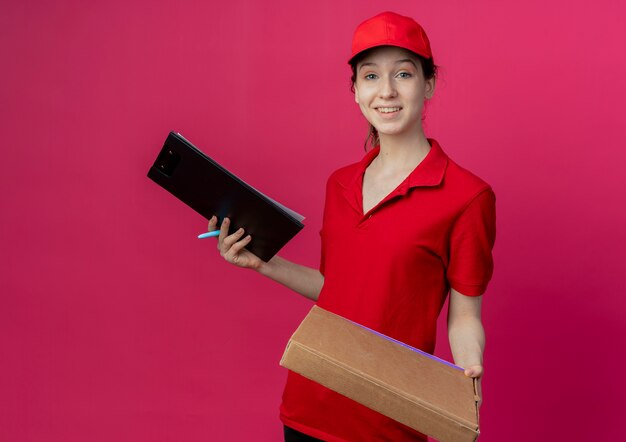 Lächelndes junges hübsches Liefermädchen in roter Uniform und Kappe, die Pizzapaketstift und Zwischenablage lokalisiert auf purpurrotem Hintergrund mit Kopienraum hält