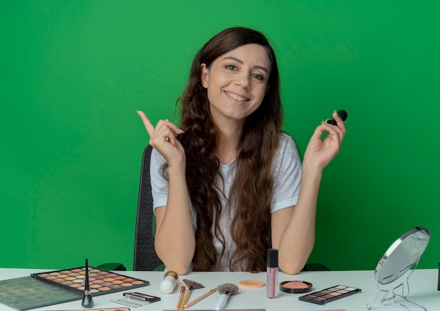 Lächelndes junges hübsches Mädchen, das am Make-up-Tisch mit Make-up-Werkzeugen hält, die Make-up-Pinsel halten und Finger lokalisiert auf grünem Hintergrund halten