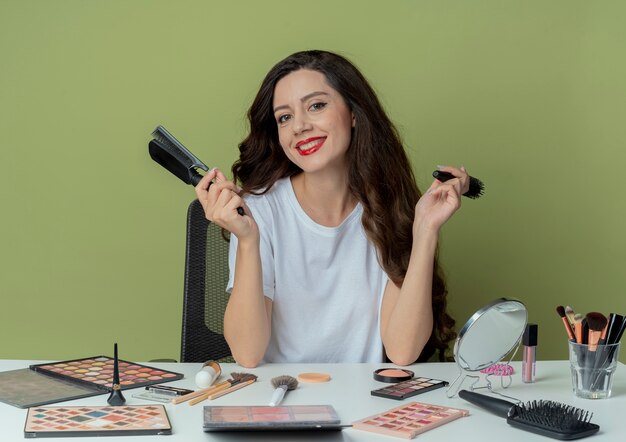 Lächelndes junges hübsches Mädchen, das am Make-up-Tisch mit Make-up-Werkzeugen sitzt, die Kämme lokalisiert auf olivgrünem Hintergrund halten