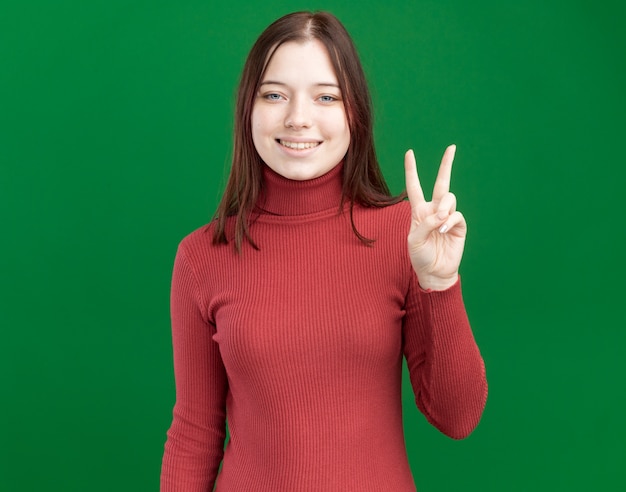 Lächelndes junges hübsches Mädchen, das Friedenszeichen lokalisiert auf grüner Wand mit Kopienraum tut