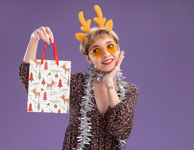 lächelndes junges hübsches Mädchen mit Rentiergeweih-Stirnband und Lametta-Girlande um den Hals mit Brille, die Weihnachtsgeschenktüte hält, die Hand auf dem Gesicht hält und die Seite isoliert auf lila Wand mit Kopienraum betrachtet