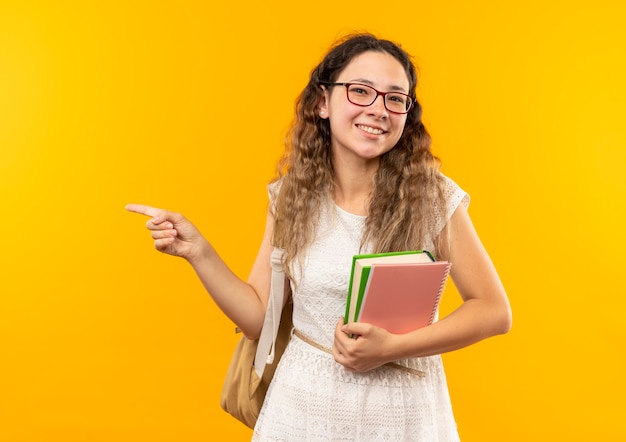 Lächelndes junges hübsches Schulmädchen, das Brille und Rückentasche hält Buch und Notizblock zeigt auf Seite lokalisiert auf gelber Wand