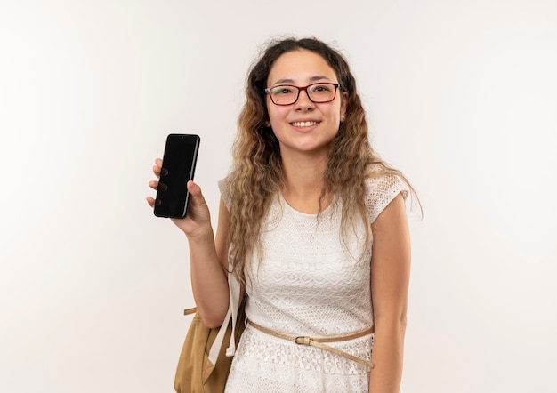 Lächelndes junges hübsches Schulmädchen, das Brille und Rückentasche trägt, die Handy lokalisiert auf weißer Wand zeigt