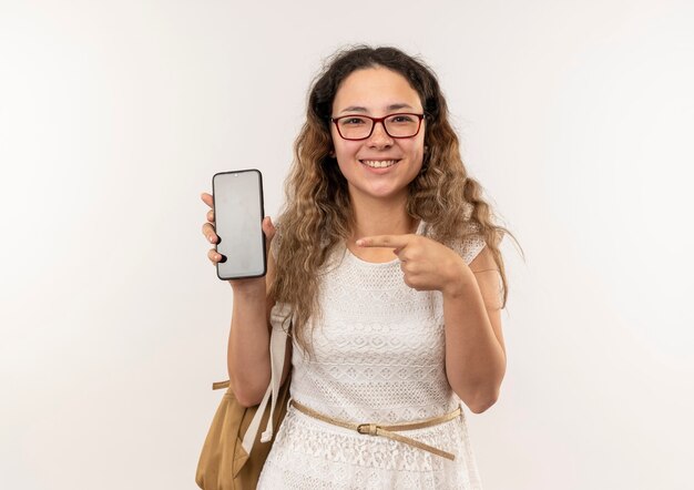 Lächelndes junges hübsches Schulmädchen, das Brille und Rückentasche trägt, zeigt und zeigt auf Handy lokalisiert auf weißer Wand