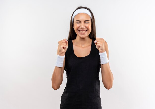 Lächelndes junges hübsches sportliches Mädchen, das Stirnband und Armband geballte Fäuste mit geschlossenen Augen auf weißem Raum trägt