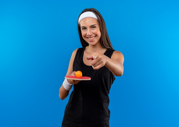 Lächelndes junges hübsches sportliches Mädchen, das Stirnband und Armband hält, das Tischtennisschläger mit Ball hält, der auf blauen Raum zeigt