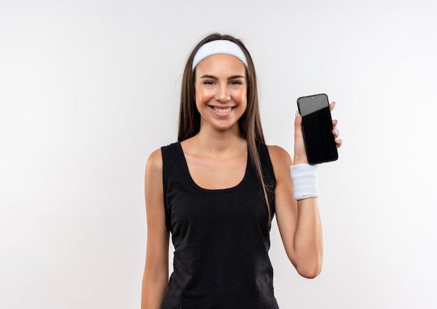 Lächelndes junges hübsches sportliches Mädchen, das Stirnband und Armband hält Handy auf weißem Raum hält