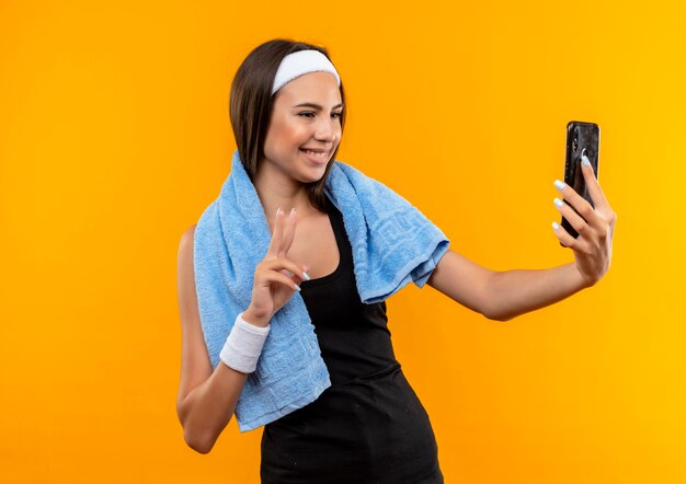 Lächelndes junges hübsches sportliches Mädchen, das Stirnband und Armband hält und Handy mit Handtuch um ihren Hals lokalisiert auf orange Raum hält