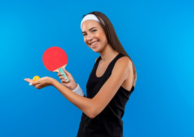 Lächelndes junges hübsches sportliches Mädchen, das Stirnband und Armband trägt, das Tischtennisschläger und Ball auf blauem Raum hält