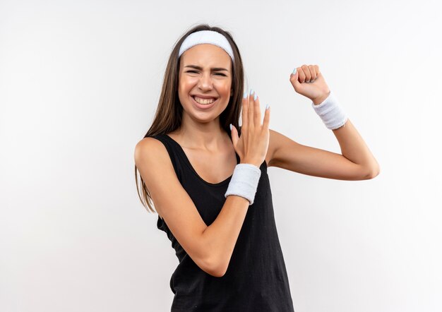 Lächelndes junges hübsches sportliches Mädchen, das Stirnband und Armband trägt und stark gestikuliert und Hand auf Leerraum erhebt