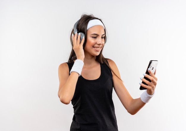 Lächelndes junges hübsches sportliches Mädchen, das Stirnband und Armband und Kopfhörer trägt, die Handy auf weißem Raum halten und betrachten