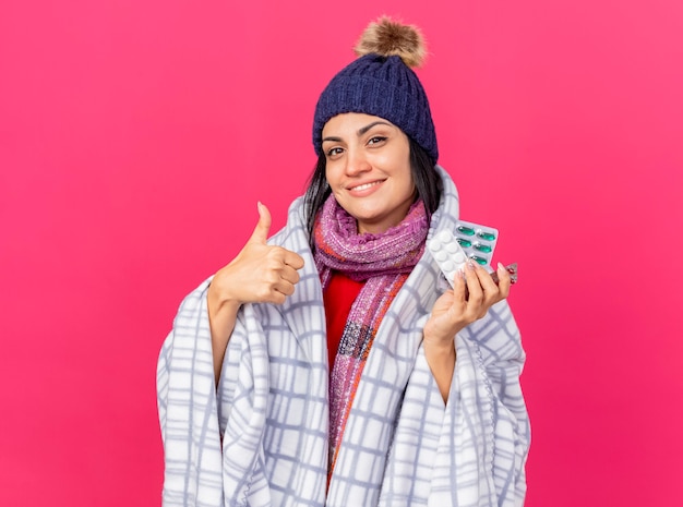 Lächelndes junges kaukasisches krankes Mädchen, das Wintermütze und Schal trägt, die in Plaid eingewickelt betrachten Kamera, die Packungen von Pillen hält, die Daumen oben lokalisiert auf purpurrotem Hintergrund mit Kopienraum zeigen