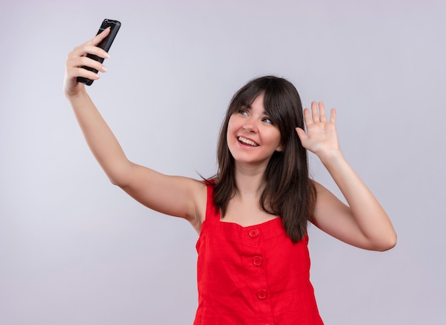 Lächelndes junges kaukasisches Mädchen, das Telefon hält und leere Hand zeigt, die Telefon auf lokalisiertem weißem Hintergrund betrachtet