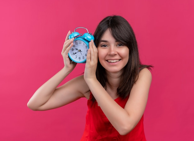 Lächelndes junges kaukasisches Mädchen, das Uhr mit beiden Zeigern hält und Kamera auf lokalisiertem rosa Hintergrund betrachtet
