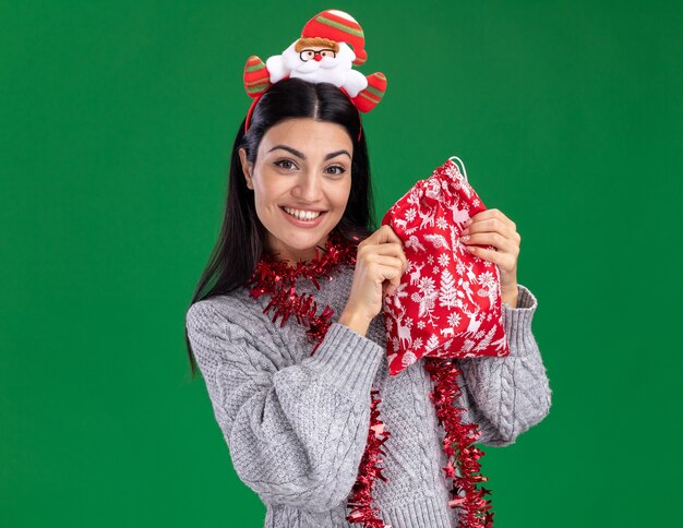 Lächelndes junges kaukasisches Mädchen, das Weihnachtsmann-Stirnband und Lametta-Girlande um den Hals hält, der Weihnachtsgeschenksack hält, der Kamera lokalisiert auf grünem Hintergrund mit Kopienraum betrachtet