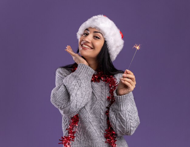 Lächelndes junges kaukasisches Mädchen, das Weihnachtsmütze und Lametta-Girlande um Hals hält, die Feiertags-Wunderkerze hält, die Kamera hält Hand unter Kinn lokalisiert auf lila Hintergrund mit Kopienraum