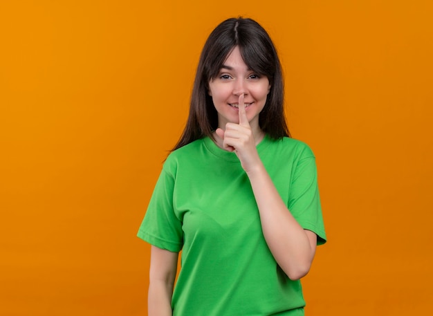 Lächelndes junges kaukasisches Mädchen im grünen Hemd setzt Finger auf Mund und betrachtet Kamera auf lokalisiertem orange Hintergrund mit Kopienraum