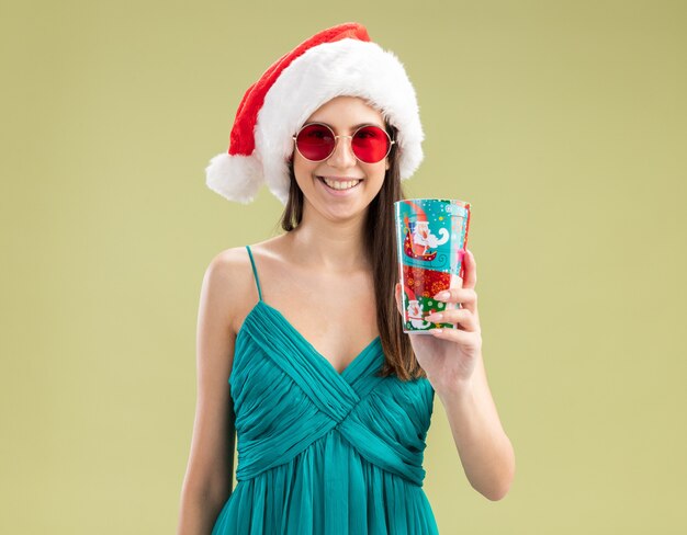 Lächelndes junges kaukasisches Mädchen in Sonnenbrille mit Weihnachtsmannhut, der Pappbecher hält