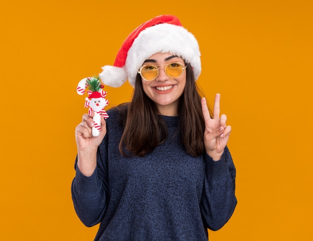 Lächelndes junges kaukasisches Mädchen in Sonnenbrille mit Weihnachtsmütze gestikuliert Victory-Zeichen und hält Zuckerstange isoliert auf oranger Wand mit Kopierraum