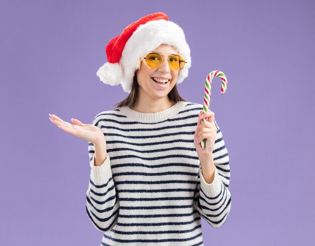 Lächelndes junges kaukasisches Mädchen in Sonnenbrille mit Weihnachtsmütze hält Zuckerstange und hält Hand offen