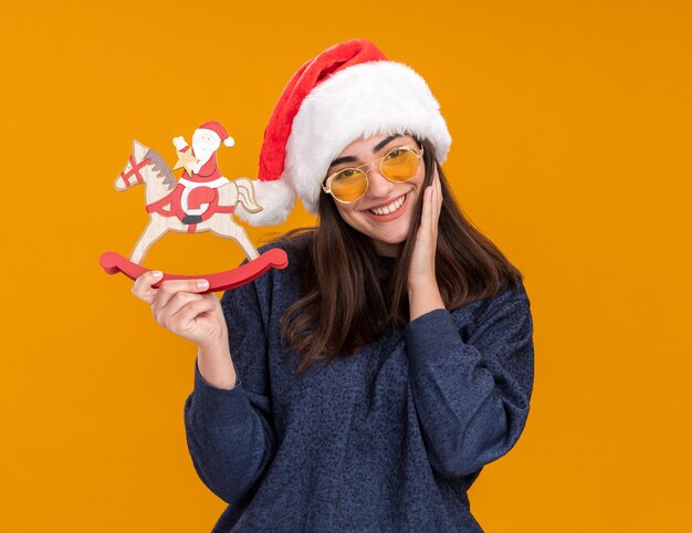 Lächelndes junges kaukasisches Mädchen in Sonnenbrille mit Weihnachtsmütze legt die Hand auf das Gesicht und hält den Weihnachtsmann auf der Schaukelpferddekoration, die auf orangefarbener Wand mit Kopierraum isoliert ist