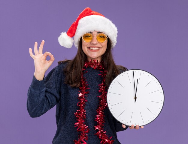 Lächelndes junges kaukasisches Mädchen in Sonnenbrille mit Weihnachtsmütze und Girlande um den Hals hält Uhr und Gesten ok Zeichen isoliert auf lila Wand mit Kopierraum