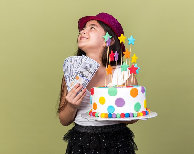 Lächelndes junges kaukasisches Mädchen mit lila Partyhut, das Geburtstagskuchen und Geld hält und auf die Seite isoliert auf olivgrüner Wand mit Kopienraum schaut