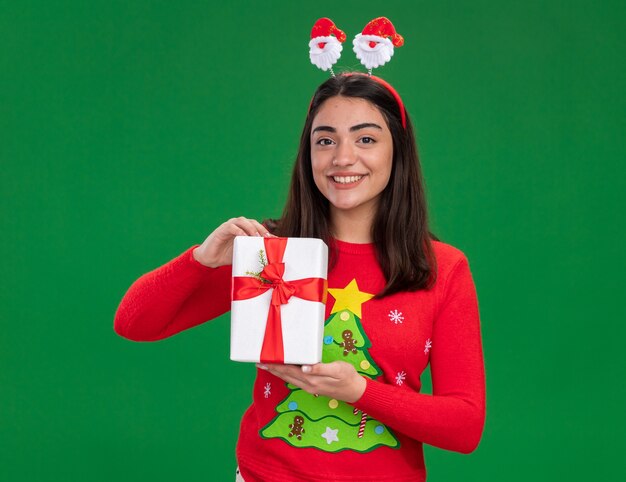 Lächelndes junges kaukasisches Mädchen mit Santa Stirnband hält Weihnachtsgeschenkbox lokalisiert auf grünem Hintergrund mit Kopienraum