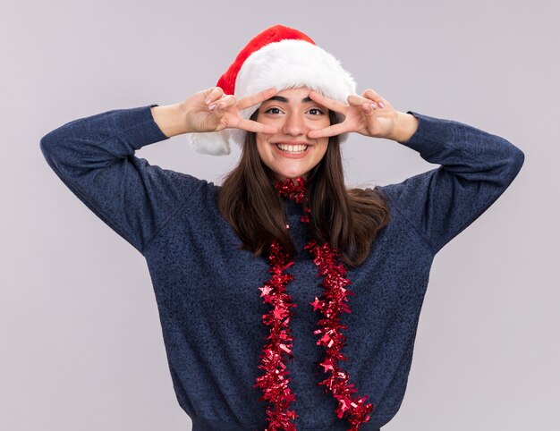 Lächelndes junges kaukasisches Mädchen mit Weihnachtsmütze und Girlande um den Hals gestikuliert Victory-Zeichen durch die Finger isoliert auf weißer Wand mit Kopierraum