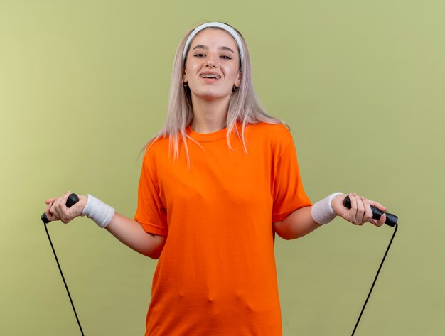 Lächelndes junges kaukasisches sportliches Mädchen mit Hosenträgern, das Stirnband und Armbänder trägt, hält Springseil