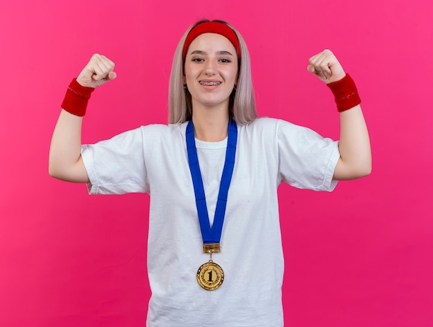 Lächelndes junges kaukasisches sportliches Mädchen mit Hosenträgern und mit Goldmedaille um den Hals, das Stirnband und Armbänder trägt, spannt Bizeps an