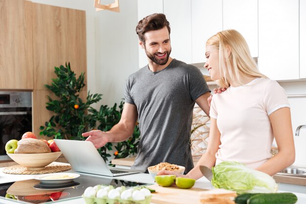 Lächelndes junges liebendes Paar, das zusammen mit Laptop kocht