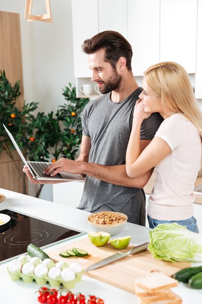 Lächelndes junges liebendes Paar, das zusammen mit Laptop kocht