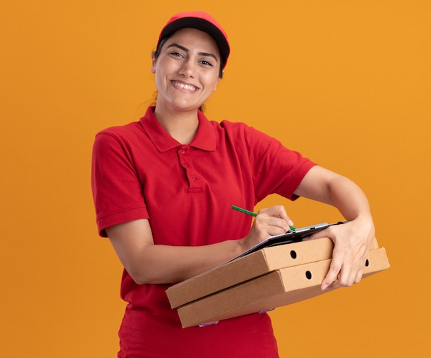 Lächelndes junges Liefermädchen, das Front betrachtet, Uniform und Mütze tragend, die etwas auf Zwischenablage auf Pizzaschachteln lokalisiert auf orange Wand schreibt