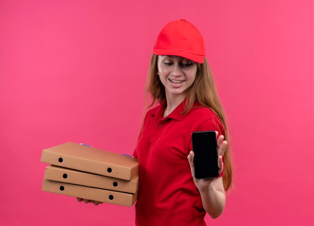 Lächelndes junges Liefermädchen, das Pakete und Handy hält und Handy auf lokalisiertem rosa Raum betrachtet