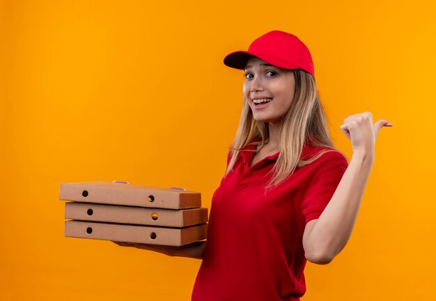 Lächelndes junges Liefermädchen, das rote Uniform und Kappe hält, die Pizzaschachtel und zur Seite lokalisiert auf orange Wand hält