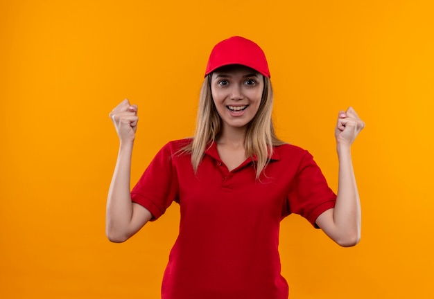 Lächelndes junges Liefermädchen, das rote Uniform und Kappe trägt, die ja Geste lokalisiert auf orange Wand zeigt