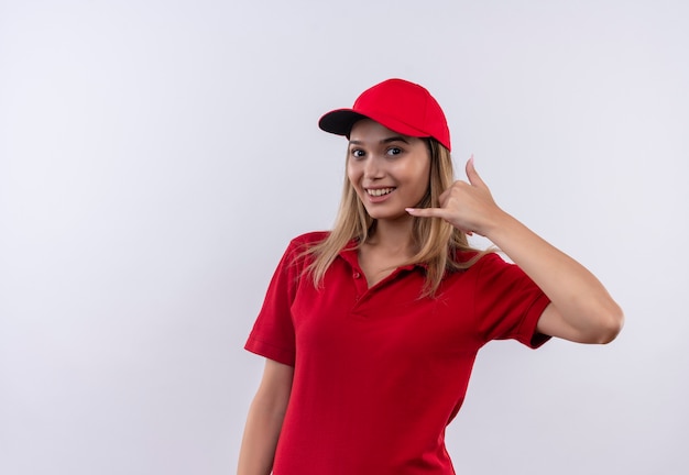 Lächelndes junges Liefermädchen, das rote Uniform und Kappe trägt, die Telefonanrufgeste lokalisiert auf Weiß zeigt