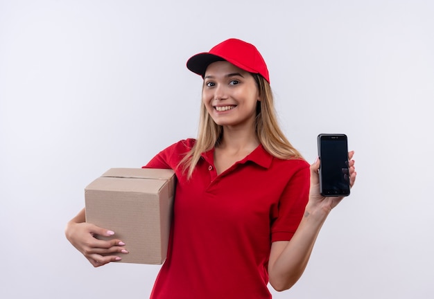 Lächelndes junges Liefermädchen, das rote Uniform und Kappenhaltebox und Telefon lokalisiert auf Weiß trägt